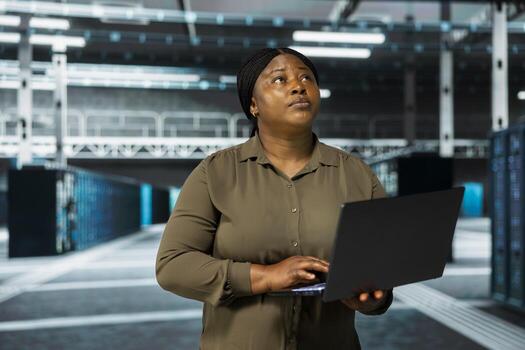 Admin using laptop to do maintenance in data center with setup helping companies stay connected. Woman holding notebook walks in server farm with machines blinking lights showing system activity photo