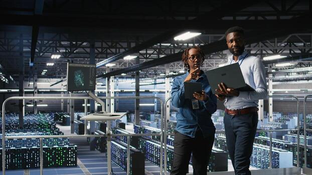 Data center coworkers doing maintenance tasks on laptop, ensuring optimal performance. Teamworking colleagues reviewing diagnostics and applying patches to server hub, improving stability, photo