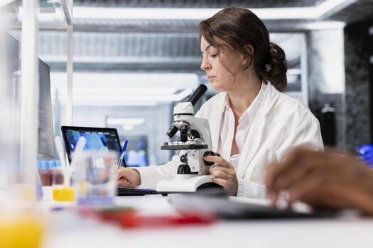 Scientist using microscope to evaluate cellular structures in lab workspace, writing down logs. Woman observes specimens through magnification tool at laboratory bench workstation, taking notes photo