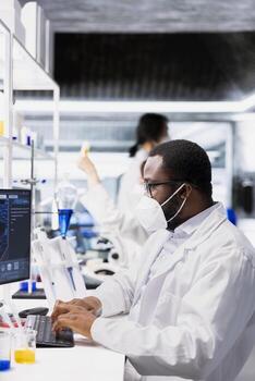 Researcher wearing medical mask using computer application for DNA data analysis in laboratory. Research facility expert wearing protective face mask using bioinformatics genomics software on PC photo