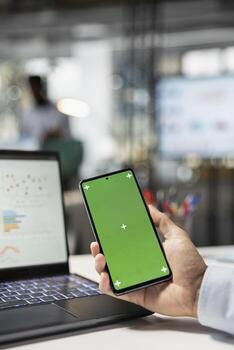 Closeup of person holding smartphone with blank mockup template. Business consultant at desk reviews company data on laptop while using green screen cellphone in modern office workspace. photo