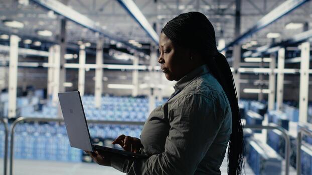 IT expert monitors server farm activity using laptop while walking in facility. African american woman in data center reviews rigs tests results to ensure systems are working correctly, camera B photo