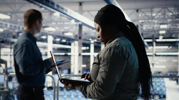 Data center admin investigates AI configurations using laptop, making adjustments to ensure optimal function. African american woman in server hub checks deep learning errors reports, camera B photo