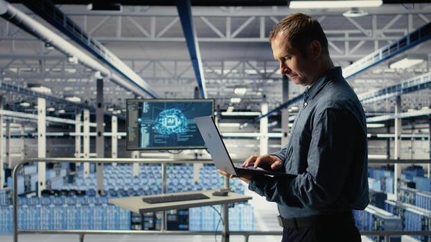 Technician monitors server room activity using laptop while walking through facility. Data center IT professional reviews rigs tests results to ensure systems are working correctly, camera A photo