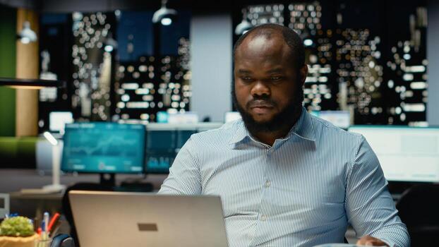 Male company analyst reading data reports and fact checking on laptop, planning project and scheduling tasks after hours. Notetaking and handwriting for objectives achievement. photo