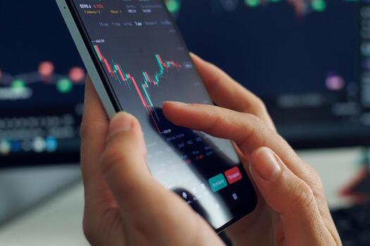 A person's hands operate a smartphone displaying a dynamic stock market chart with fluctuating red and green bars, against a blurred background of a larger monitor showing similar financial data. photo