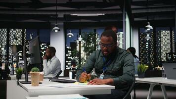 Black business person placing a sticky note reminder on monitor, using adhesive paper label to remember a new development task. Dark office at night, motivation for efficiency. Camera A. video