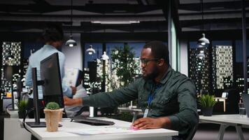 afrikanisch amerikanisch Berater extrahieren Daten durch Hand auf ein Notizbuch spät beim Nacht, Abfassung Highlights von Finanzen Berichte zum das Geschäft Verwaltung. Notizen unterstützt Tor Einstellung. Kamera a. video