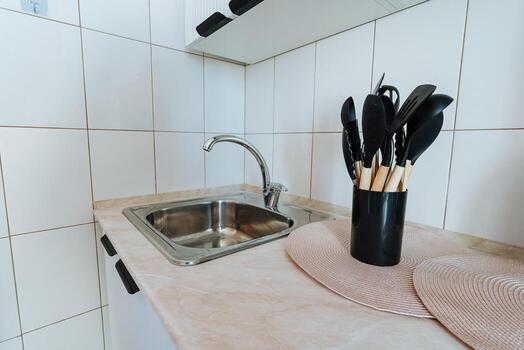 In the kitchen area, there is a sink installed with several utensils placed inside a stylish black cup that sits neatly on the countertop, highlighting the rooms design and functionality photo