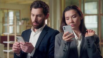 Multiethnic business colleagues using devices at office Korean businesswoman scrolling mobile phone thoughtful envious glancing coworker screen Caucasian man checks online networking browsing internet video