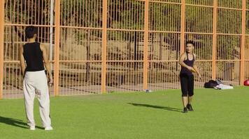 man framställning kasta med gul frisbee medan partner väntar på grön artificiell torva, metall staket och träd bakgrund, dynamisk verkan ögonblick. video