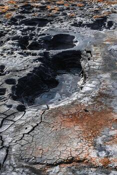 Close view of a geothermal mud field in Iceland, with cracked clay, dark viscous pools, rust hued deposits, and sulfur stains under diffused daylight, emphasizing volcanic activity. photo
