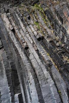 Steep basalt columns with sharp hexagonal edges rise on a cliff in Iceland. Natural directional light reveals mineral lines and sparse plants in tight composition. photo