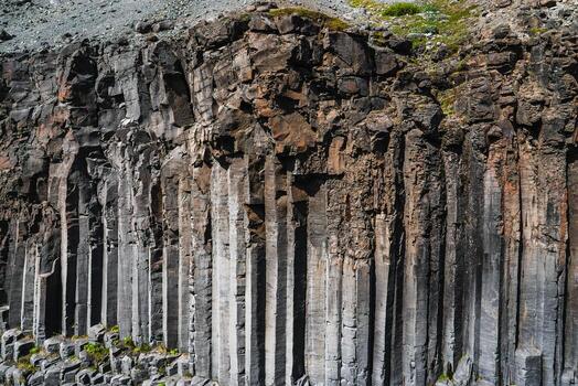 Hexagonal basalt columns form vertical stacks along a rugged cliff in Iceland. Midday light reveals fractured textures and gray tones, with patches of green moss. photo