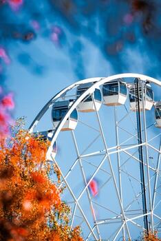 A white Ferris wheel in Riga rises above orange leaves at midday, angular gondolas gleam in clear blue light, upward angle and shallow depth of field create a crisp look. photo