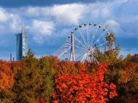 Aerial view over Riga, Latvia shows a Ferris wheel above orange and red trees, with Vansu Bridge and Z Towers visible on a clear fall afternoon with dramatic clouds. photo