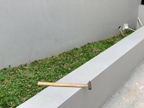 A simple wooden hammer rests on a low concrete wall, with green grass and a plain gray wall in the background, creating a minimalist and functional scene. photo