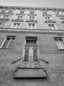 Upward view of old European building facade with ornate balcony door and symmetrical window layout. Captured in black and white with strong vertical perspective. Suitable for themes of architecture photo
