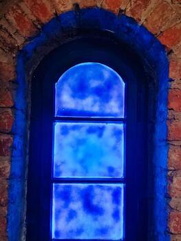 Arched window with frosted glowing blue glass, surrounded by old red brick wall. Close-up view in artificial lighting photo