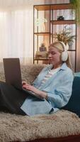 jovem mulher goza freelance trabalhos às lar, digitando em computador portátil com calma foco ouvindo quieto música em fones de ouvido. menina em sofá descansa, caloroso pôr do sol, Misturando conforto, profissional e criativo comprometimento video