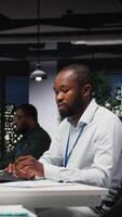 Vertical African american male worker proofreading financial numbers, reviewing archived files after hours in the strategy room. Man evaluating data on reports and doing research. Camera A. video