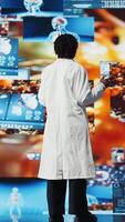 vertikal sjukvård expert- uppslukad förbi trogen ct skannar, mri data, och labb analys visas på videovägg. forskare i medicinsk forskning labb förvånad förbi hälsa data på hög tech paneler, kamera en video