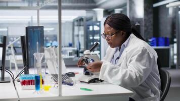 Researcher using microscope and specimen tray in laboratory workspace. Optical instrument, lens system and magnifying glass support clinical testing and biotechnology for healthcare. Camera B. video