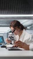 vertikal cytoteknolog justerar mikroskop, zoomar på cellulär strukturer i labb, studier genetisk störningar. molekyl biolog konstaterar mikroorganismer med förstoring verktyg på laboratorium bänk, kamera en video