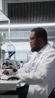 Vertical Clinical microbiologist using compound microscope for detailed sample observation using PC software. Biotechnology research facility worker studies tissue with optical device and computer, camera A video