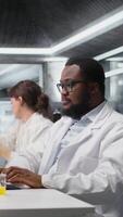 Vertical Portrait of smiling laboratory scientist using computer monitor, processing DNA patient data for clinical research. Cheerful african american lab worker looking at analysis diagnostics on PC, camera A video
