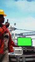 Vertical Isolated screen laptop used by drilling rig crew colleagues holding radio device to monitor systems. Engineering team on offshore platform deck using walkie talkie and chroma key notebook, camera A video