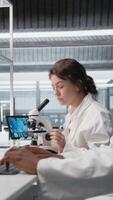 Vertical Zoom in on histology technician in lab using scientific microscope for cellular analysis. Woman studies microorganisms inside modern research facility with optical device equipment, camera A video