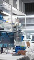 Vertical High tech lab technician at desk typing on PC keyboard, looking at molecular model, doing research tasks. African american man using bioengineering program on PC in medical lab, camera A video
