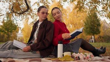 två universitet studenter sitta tillbaka till tillbaka på picknick filt i campus parkera, jämförande anteckningar och läsning kapitel medan gyllene löv mjukt falla runt om dem och termos med muggar står förbi för värma studie ha sönder. video