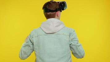 bak- se av förvånad tonåring studerande pojke använder sig av headsetet hjälm app spela simulering spel. tittar på virtuell verklighet 3d 360 . ung man i vr glasögon isolerat på gul bakgrund. framtida teknologi video