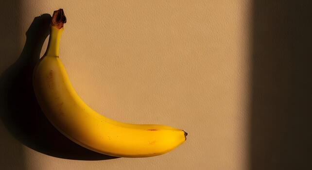 Minimalist top view of a single ripe yellow banana with a dramatic shadow from direct sunlight on a warm, neutral background photo