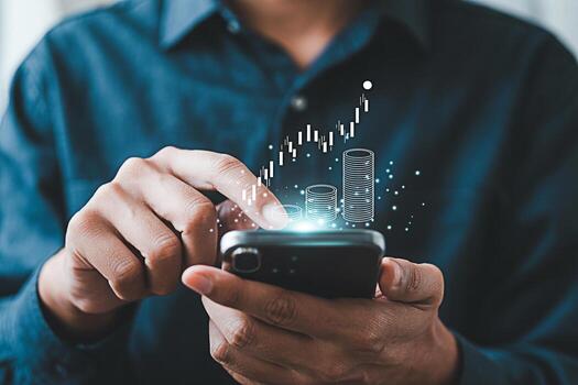 Man in a blue shirt interacts with a smartphone displaying a rising stock chart and coin stacks symbolizing financial growth and investment success in the digital age creating a sense of optimism and photo