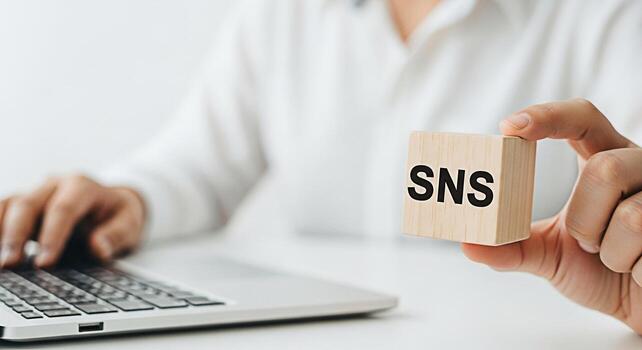 Person typing on a laptop while holding a wooden block with the letters SNS in a bright minimalist office environment conveying a message about social networking services and online communication photo