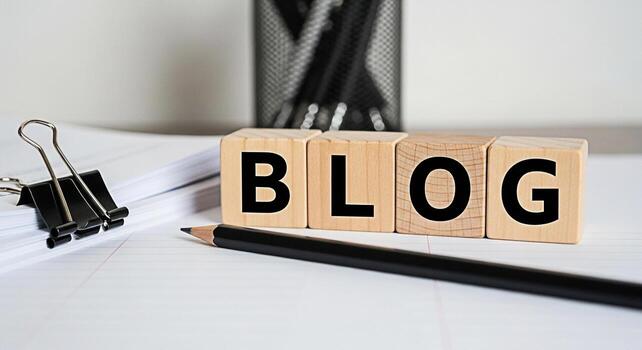 Wooden blocks spelling BLOG on a lined paper surface accompanied by a pencil and paper stack suggesting content creation and online writing in a bright organized workspace fostering a creative and pro photo