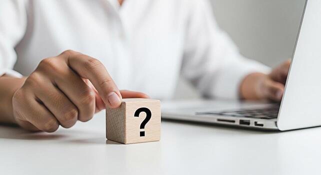 Persons hand touching a wooden block with a question mark while working on a laptop in a bright modern office representing uncertainty problemsolving and the search for answers with technology photo