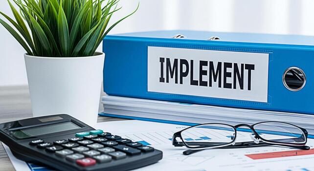 Office desk setup with a blue binder labeled IMPLEMENT calculator glasses and financial charts conveying a sense of organization focus and the execution of business strategies in a professional settin photo
