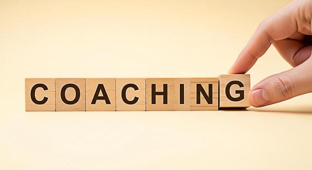 A hand arranging wooden blocks spelling COACHING on a light yellow surface symbolizing guidance and support in a learning environment fostering a positive and encouraging atmosphere for personal and p photo