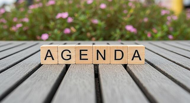 Wooden blocks spelling AGENDA displayed on a gray wooden table with a blurred background of pink flowers in a garden setting representing organization planning and a structured approach to tasks and m photo