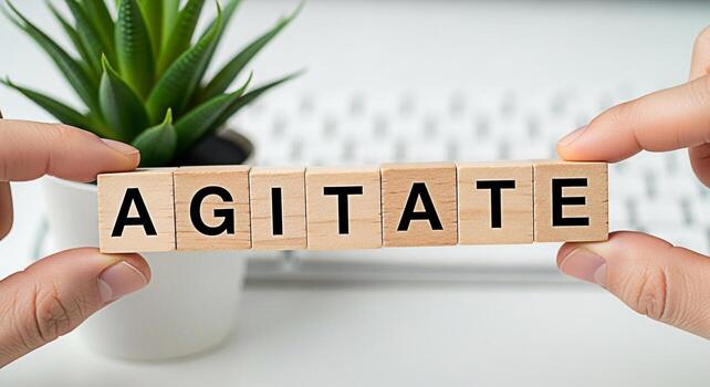 Hands holding wooden blocks spelling AGITATE in a bright office setting symbolizing stirring up emotions and creating a sense of urgency and unrest ideal for illustrating conflict or debate photo