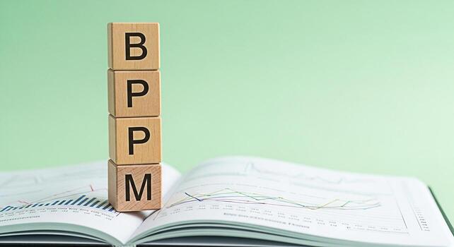 Wooden blocks spelling BPM stacked on an open book with financial charts against a light green background representing business process management and efficiency in a corporate environment conveying a photo