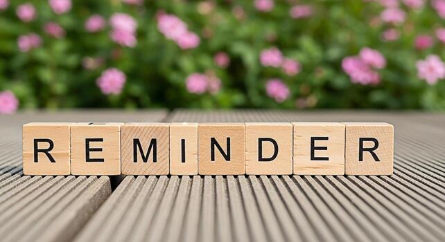 Wooden blocks spelling REMINDER on a wooden surface with a blurred background of pink flowers serving as a gentle nudge and a visual cue for memory and important tasks in a serene outdoor setting photo