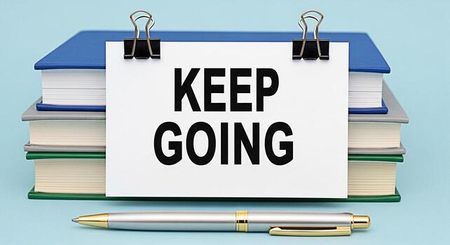 Stack of colorful books displaying a motivational message Keep Going on a white card with binder clips alongside a sleek silver pen on a light blue surface conveying a message of perseverance and enco photo