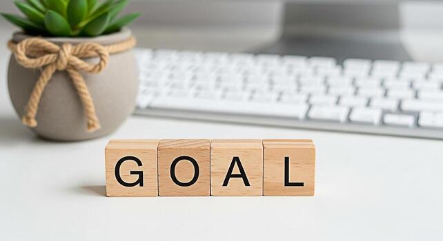 Wooden blocks spelling GOAL on a bright white desk with a potted succulent and keyboard in the background representing ambition achievement and focus in a modern workspace environment photo