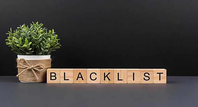 Wooden blocks spelling Blacklist arranged on a dark surface next to a potted plant symbolizing exclusion and restriction in a business or personal context creating a somber and cautionary mood photo