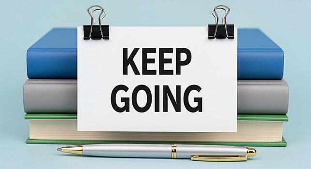 Stack of colorful books displaying a motivational message Keep Going on a white card with binder clips placed on a light blue surface symbolizing perseverance and encouragement in education photo